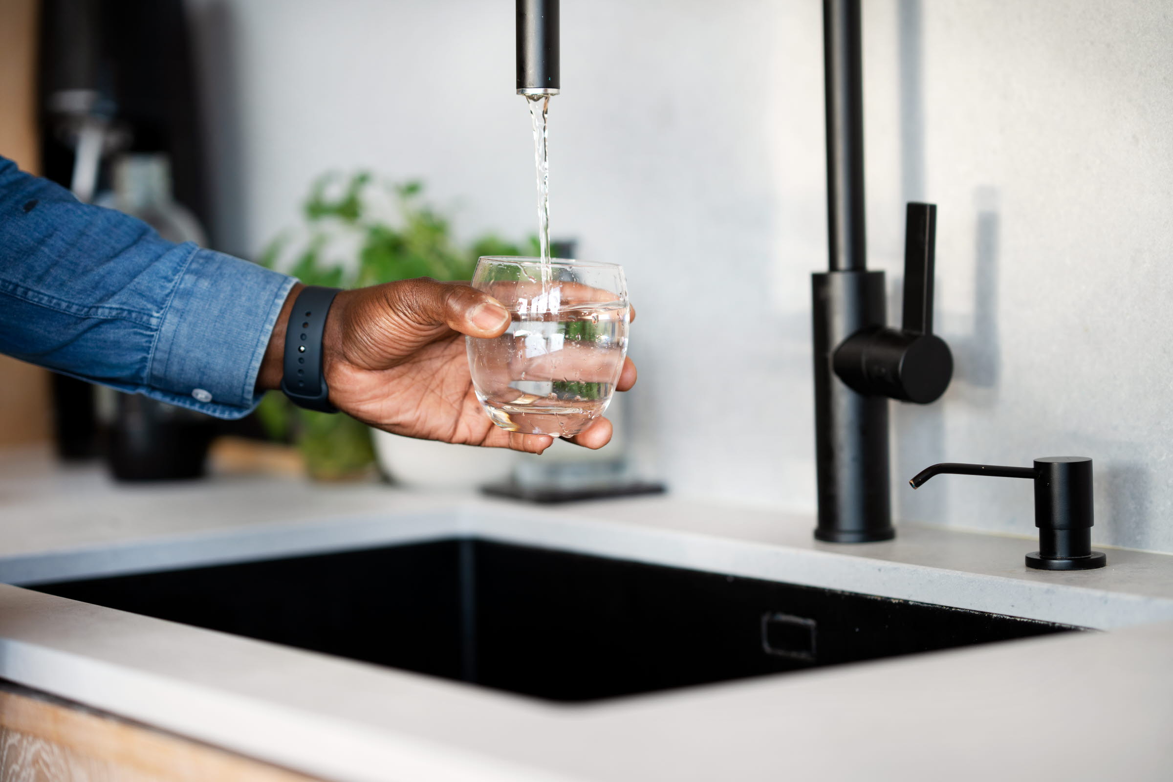Ett kök med en ljus diskbänk med svart ho och kökskran, där en hand fyller på ett glas vatten.